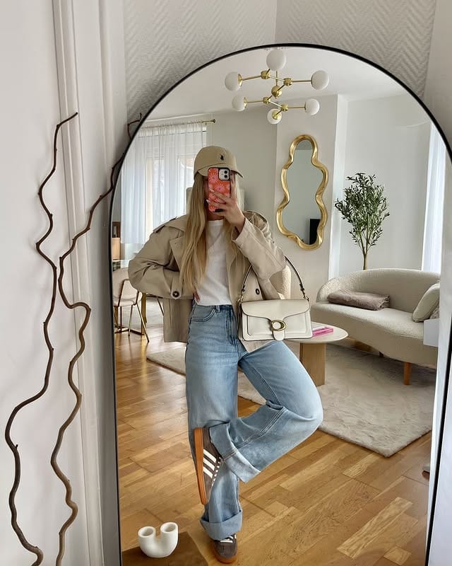 Casual fashion with beige jacket, wide leg jeans, sporty sneakers and matching bag accessory, reflected in a decorative mirror in the stylish living room with modern furniture and plants.