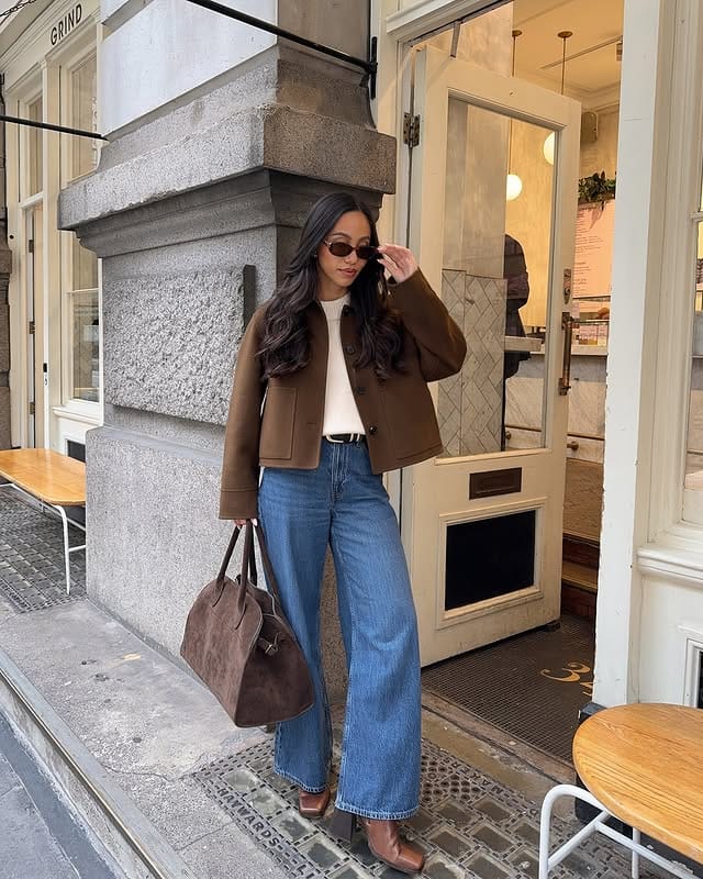 Stylish woman with long hair, wearing sunglasses, a brown jacket, a white blouse and loose jeans, carrying a shoulder bag, ready for a day out. casual outfit