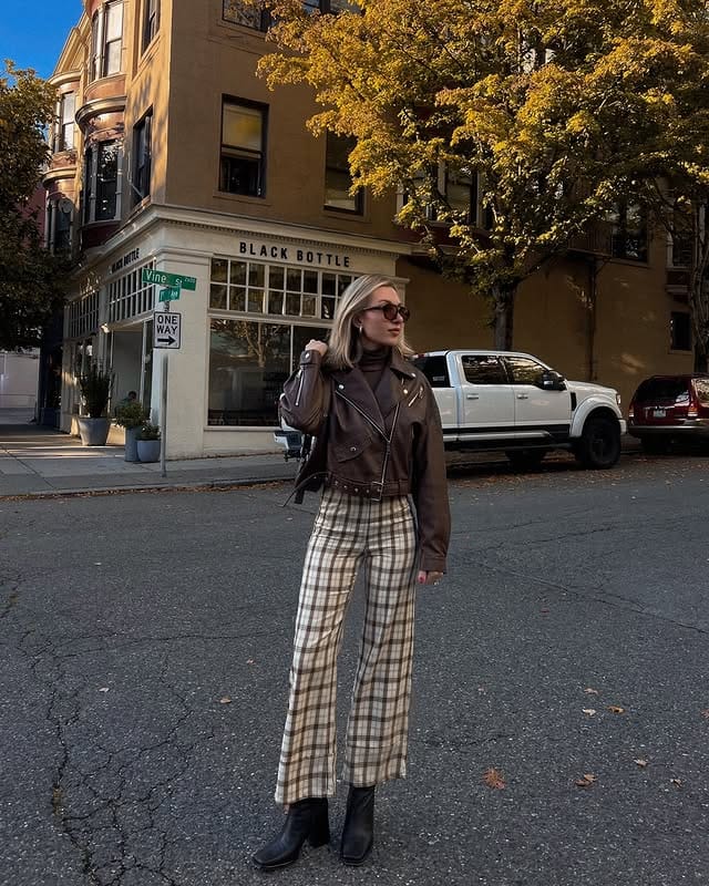 Woman wearing a leather jacket, plaid jeans, and black boots walking down the street on an autumn day, with colorful trees and old buildings in the background, promoting urban fashion and casual style