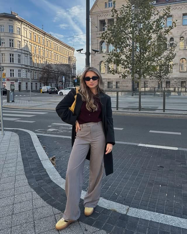 Modern woman with sunglasses, black blazer, wine-colored blouse, beige wide-leg pants, and yellow sneakers, posing on the street with historic buildings in the background, reflecting urban and contemporary style.