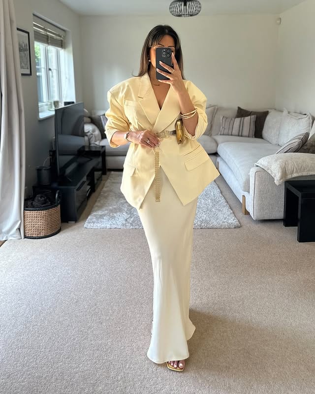 Image of an elegant woman wearing a beige blazer and a long skirt, taking a selfie in the mirror of a modern living room with comfortable furniture and clean decor.