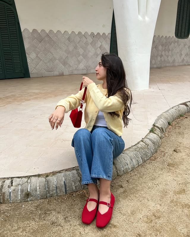Young woman sitting next to a tree, with long dark hair, wearing a yellow jacket, jeans and red shoes, in an urban space with a decorative wall in the background.