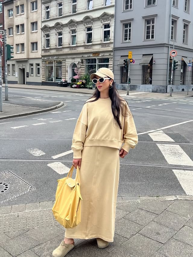 Stylish woman in light-colored clothing and modern accessories crossing a street in a city center, highlighting her yellow bag and sunglasses.