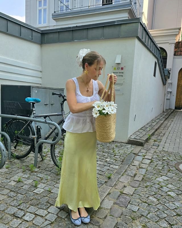 Woman wearing a long yellow skirt and white blouse, holding a basket with white flowers on the street, next to bicycles and modern buildings.