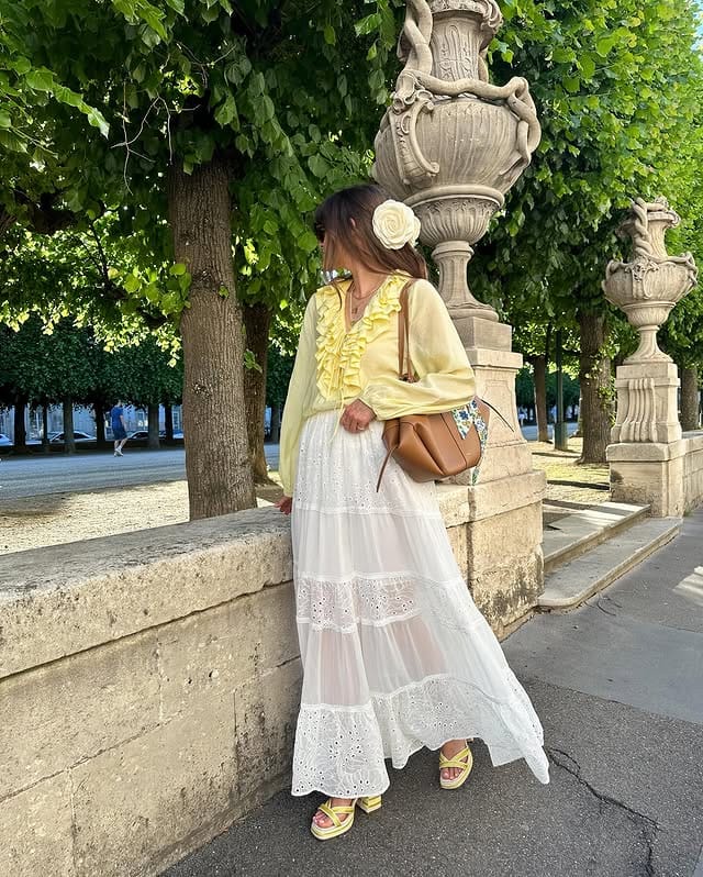 Elegant woman in long white dress and butter yellow blouse, wearing sandals and carrying a brown bag, posing next to a fountain in a park, surrounded by green trees and natural light.