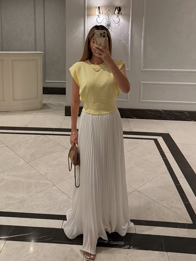 Image of a woman wearing a white dress and yellow blouse, taking a selfie in an elegant setting with marble details and sophisticated lighting.