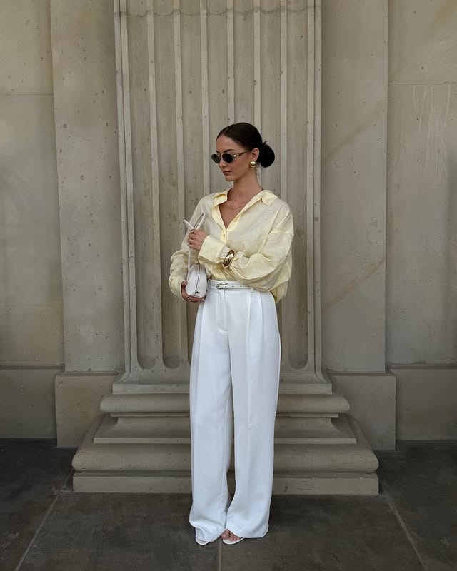 Elegant woman wearing sunglasses, yellow silk blouse and white pants, posing against a stone wall, sophisticated and fashionable style.