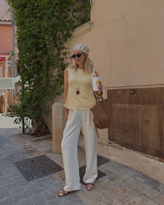 Modern woman dressed in light clothing, hat, sunglasses and carrying a drink on a sunny street, urban and relaxed environment.