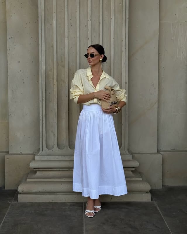 Elegant woman wearing sunglasses, a yellow shirt and a white skirt, holding a paper bag, posing next to a stone wall, with a sophisticated and modern style.