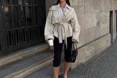 Elegant woman with sunglasses, beige cropped trench coat with a belt at the waist, black capri pants and heeled sandals in an urban setting.